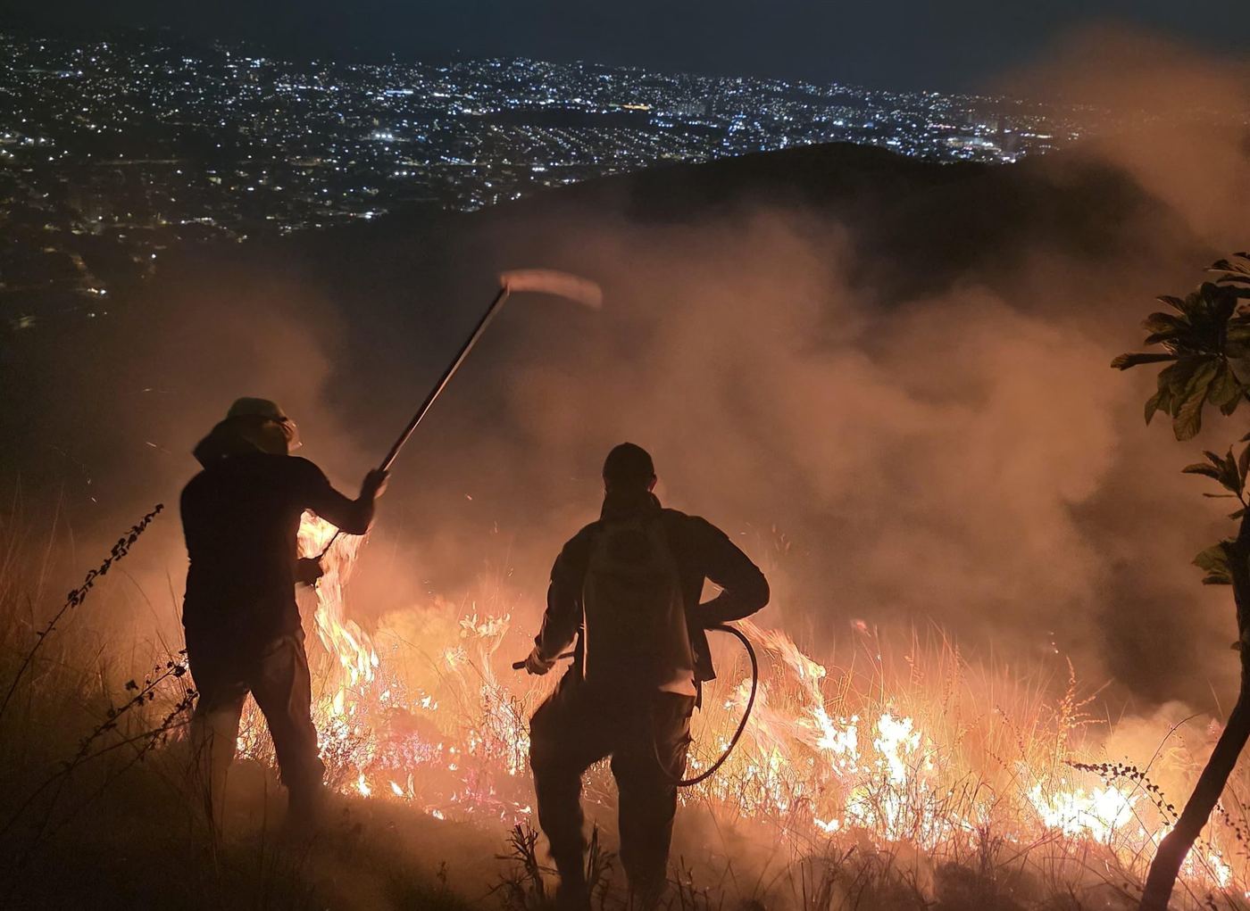Two volunteer firefighters facing flames at night, with the city lights behind